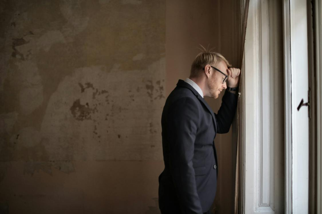 A desperate male entrepreneur standing near a window, symbolizing the emotional toll and challenges associated with fear of failure and setbacks in business.