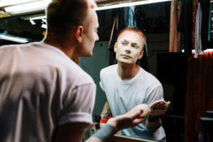 A person with dramatic makeup in progress, looking into a mirror and applying lipstick.