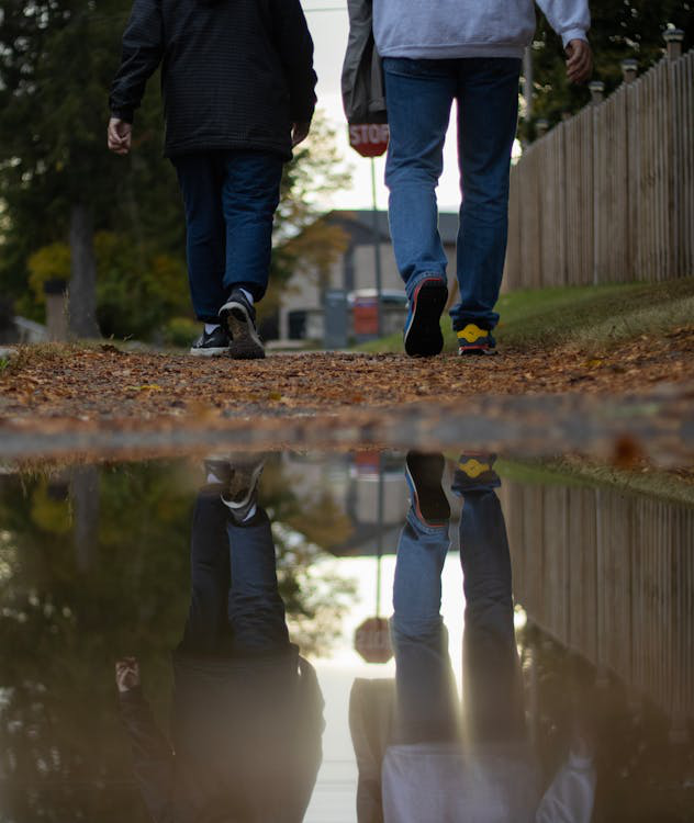 People walking on a sidewalk, with their reflections distorted in a puddle of water.