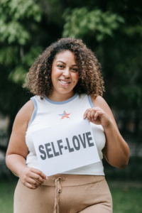 A woman holding a white piece of paper with the words "Self Love" written on it in black.
