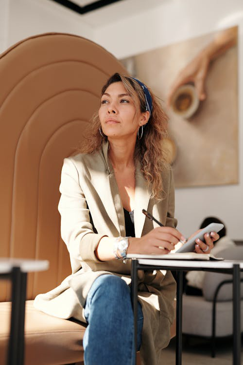 A woman wearing a blazer and blue denim jeans is sitting in a café writing in her journal