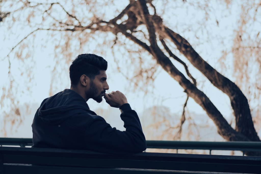 A man in a black hoodie sitting on a bench near green trees